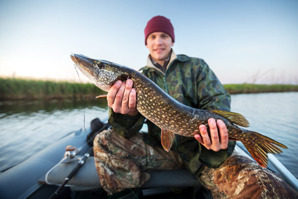 Happy young fisherman holding pike and sitting in the boat on the lake